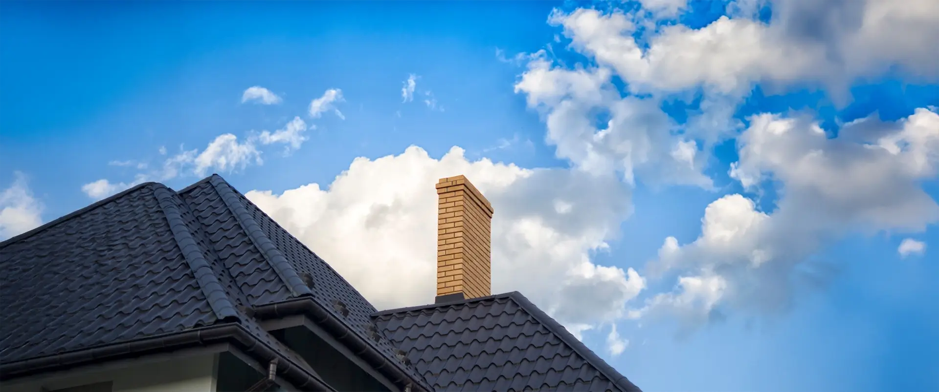 Chimney made out of bricks on top of a ceramic tile roof. We see a blue sky with white clouds in the background.
