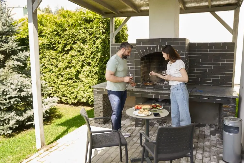 A couple cooks together in a backyard kitchen featuring a chimney-style grill.