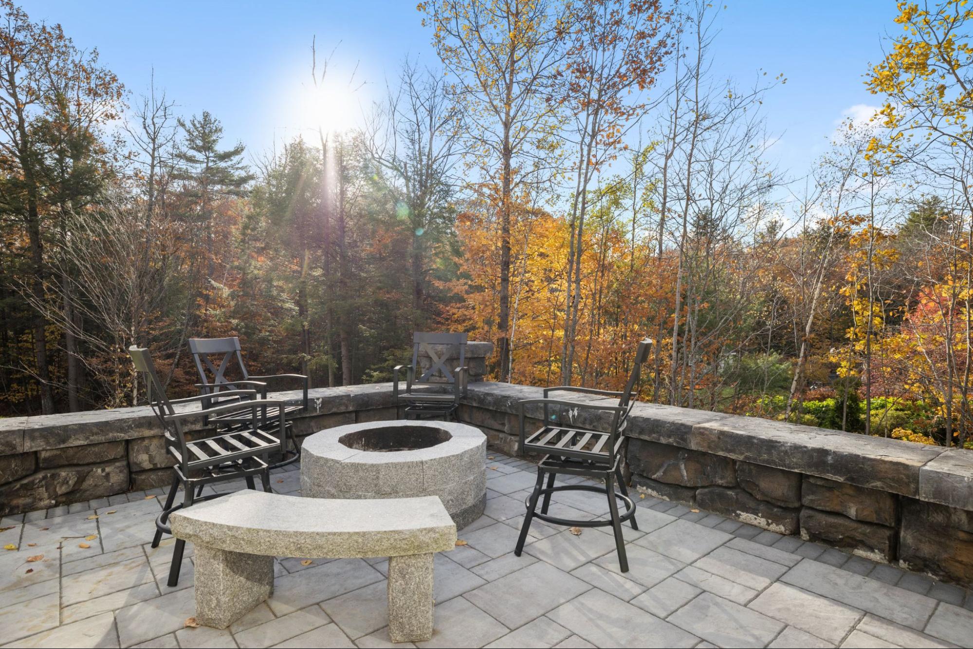 A back stone work patio with steel chairs and a fire pit set in front of tall trees.